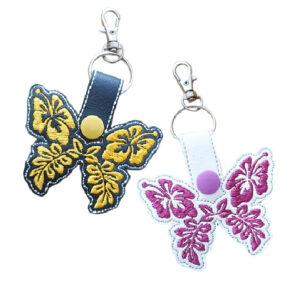 Two embroidered hibiscus butterfly keyrings made from faux leather, one black with yellow floral embroidery and one white with pink floral embroidery, each attached to a metal keyring and lobster clasp on a white background.