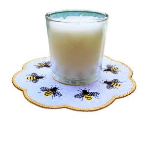 Glass jar candle placed on a flower-shaped Honey Bee candle mat with embroidered yellow and black bees, a subtle honeycomb stitched background, and a golden scalloped border.