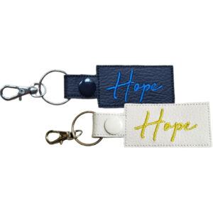 Two rectangular keyrings with the word "Hope" embroidered in cursive. One keyring is black with blue embroidery and a silver clasp, and the other is white with yellow embroidery and a silver clasp.