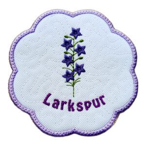 Round scalloped candle coaster embroidered with purple larkspur flowers and green stems, featuring a subtle textured background, a purple stitched border, and the word “Larkspur” embroidered beneath the design.