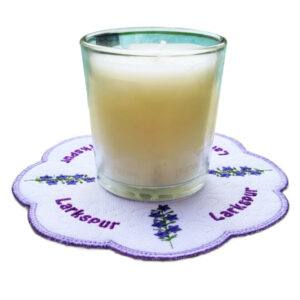 Glass candle placed on a scalloped embroidered larkspur candle mat, featuring purple larkspur flowers with green stems, a subtle textured background, and a purple stitched border.