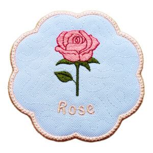 Embroidered candle coaster with a scalloped edge, featuring a pink rose with green leaves and the word “Rose” stitched below on a white background.