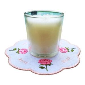 Glass candle placed on a scalloped embroidered rose candle mat, featuring pink rose motifs with green leaves, a subtle textured background, and a pale blush stitched border.