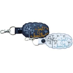 Two faux leather dandelion keyrings shown side by side. One is black with an embroidered dandelion design and the words “just breathe” stitched in yellow, and the other is white with an embroidered dandelion design and the words “just breathe” stitched in teal. Both keyrings have stitched edges, a strap with a snap button, and silver-toned lobster clasps with split rings.