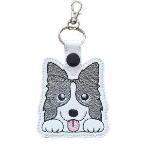 A white faux leather keyring featuring a kawaii-style embroidered Border Collie face with black and white fur, bright eyes, and a pink tongue. The keyring includes a silver split ring, swivel clasp, and black snap fastener, designed as a cute accessory for dog lovers.