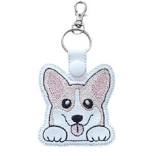 An adorable Kawaii Corgi keyring featuring a smiling corgi face with perky ears, big sparkling eyes, and a little tongue sticking out. The embroidered design is stitched on white faux leather with a white snap fastener, attached to a silver keyring and swivel clasp. A cute and cheerful accessory for dog and Corgi lovers.