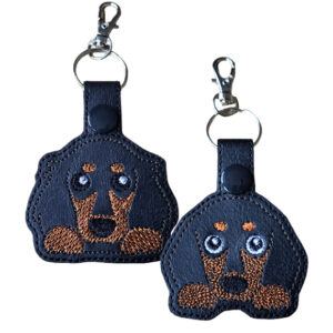 Kawaii Dachshund Keyring featuring two embroidered sausage dog designs on black faux leather – one short-haired and one long-haired, both with brown stitched details and a silver keyring clasp.