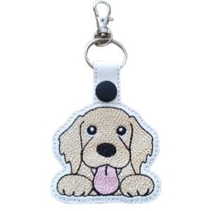 A cute embroidered keyring featuring a kawaii-style Golden Retriever face with floppy ears and a pink tongue sticking out, stitched onto white faux leather. The keyring includes a silver split ring, lobster clasp, and black snap fastener.