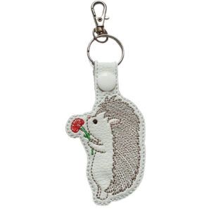An embroidered keyring shaped like a cute cartoon hedgehog standing on its hind legs and holding a red flower. The hedgehog has a light brown body, a white face and belly, and a silver ring with a clasp attached at the top.