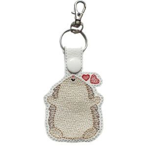 An embroidered keyring shaped like a cute cartoon hedgehog with two small red hearts above its head. The hedgehog has a light brown and cream body, a tiny stitched nose, and a silver keyring with a clasp attached at the top.