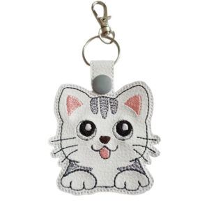 A cute embroidered keyring shaped like a cartoon kitten with big shiny eyes, pink ears, and a little tongue sticking out. The kitten has grey stripes on its head and paws, and the keyring includes a silver ring and clasp for attaching to keys or bags.