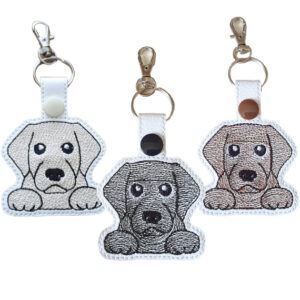 Three kawaii Labrador keyrings made from white faux leather with embroidered faces in golden, black, and chocolate brown thread. Each keyring features a secure metal ring and lobster clasp with a coordinating snap fastener, showing the playful and lovable expressions of Labradors.