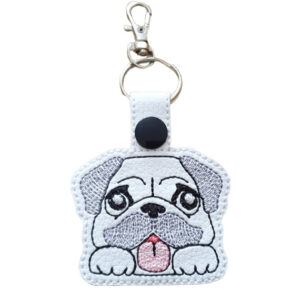 An adorable Kawaii Pug keyring featuring a cute embroidered pug face with big round eyes, floppy ears, and a pink tongue sticking out. The design is stitched on white faux leather with a black snap fastener, attached to a silver keyring and swivel clasp. A fun and charming accessory for pug fans and dog lovers.