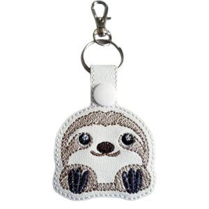 A cute embroidered keyring shaped like a cartoon sloth with big shiny eyes, a small nose, and a gentle smile. The sloth is light brown and cream with dark brown paws, and the keyring has a silver ring and clasp attached at the top.