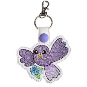 An embroidered keyring shaped like a cute cartoon bird with large eyes, a small yellow beak, and outstretched wings. The bird is holding a small blue flower, and the keyring has a silver ring and clasp attached at the top.