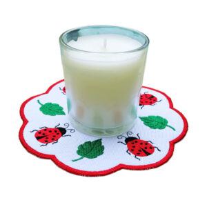 A white scalloped-edge candle mat with red trim, embroidered with red ladybirds and green leaves, shown with a glass candle placed on top to demonstrate its decorative and practical use.