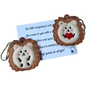 Two Little Hedgehog Pocket Pal keyrings made of soft felt, one holding a red heart and one without. Each has a ball chain for attaching to keys or bags, shown with a keepsake card featuring an uplifting message.