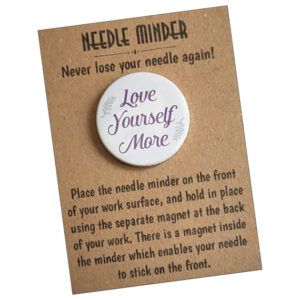 Love Yourself More Needle Minder – circular magnetic sewing accessory featuring inspirational text in elegant purple lettering with leaf details. Shown on brown kraft card packaging.