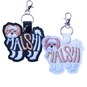 Two embroidered Malshi word art keyrings made from faux leather, showing the Malshi dog silhouette with the word “Malshi” stitched into the body. One keyring is black with light and tan stitching, and the other is white with soft tan stitching, both attached to a metal split ring and lobster clasp.