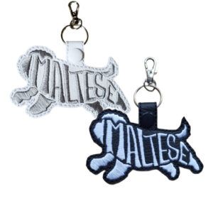Two embroidered Maltese word art keyrings made from faux leather, showing the Maltese dog silhouette with the word “Maltese” stitched into the body. One keyring is white with soft grey stitching, and the other is black with light stitching, both finished with a snap tab, metal split ring, and lobster clasp.