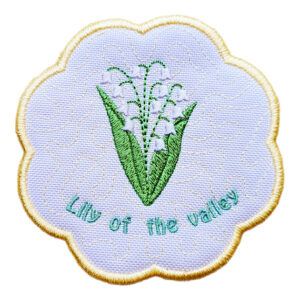 Round scalloped candle coaster with embroidered lily of the valley flowers in white and green, featuring light stippled background stitching, a pale yellow border, and the words “Lily of the valley” stitched in soft blue along the bottom.