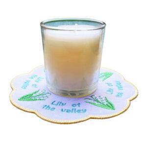 Glass candle placed on a scalloped embroidered Lily of the Valley candle mat, featuring green leaves, white bell-shaped flowers, and a pale yellow stitched border with “Lily of the valley” embroidered around the design.