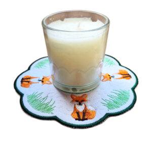 Embroidered woodland fox candle mat with scalloped edge, green border, and candle placed on top, featuring multiple fox motifs on a white background