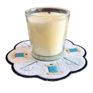 Embroidered needle and thread candle mat with scalloped edge, featuring spools of thread and stitched details on a white background, holding a glass candle on top.