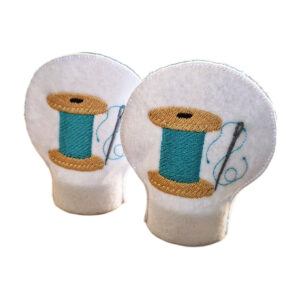 Set of two embroidered needle and thread tealight covers made from white felt, each featuring a spool of thread and stitched detail, designed to fit over LED tealights.