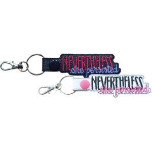 Rectangular keyring embroidered with the phrase “Nevertheless, she persisted” in two colours, attached to a metal split ring with a swivel clasp. The keyring is designed as a motivational accessory that can be clipped to keys, bags, or backpacks