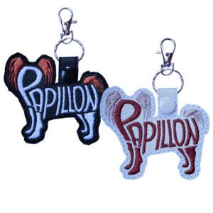 Two embroidered Papillon word art keyrings made from faux leather, showing the Papillon dog silhouette with the word “Papillon” stitched into the body. One keyring is black with light and warm orange stitching, and the other is white with warm rust and soft neutral stitching, both finished with a snap tab, metal split ring, and lobster clasp.