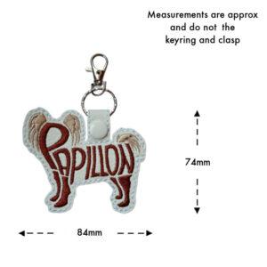 Embroidered Papillon word art keyring made from faux leather, shown with measurement guide. The Papillon dog silhouette features the word “Papillon” stitched into the body, with approximate dimensions of 84mm wide and 74mm high, not including the keyring and clasp.