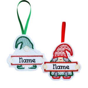 Two embroidered felt Christmas gnome ornaments with hanging ribbons. The male gnome wears a green hat with a green ribbon hanger, and the female gnome wears a red patterned hat with a red ribbon hanger. Both hold blank banners personalised with embroidered names.