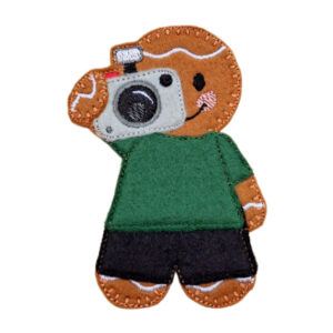 Felt gingerbread photographer character wearing a green top and black trousers, holding a stitched camera up to their face with a smiling expression.