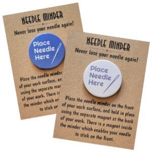 Two round needle minders displayed on kraft card backing. Each has the text ‘Place Needle Here’ with a simple needle illustration. One has a blue background with white text, and the other has a white background with purple text. The packaging reads ‘Needle Minder – Never lose your needle again!’ along with usage instructions.