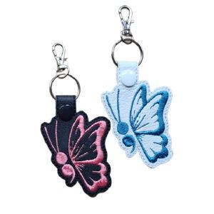 Two embroidered butterfly keyrings made from faux leather, one black with pink stitching and one white with teal stitching, each attached to a metal keyring and lobster clasp on a white background.