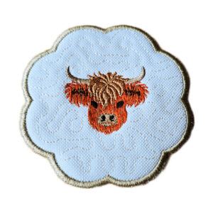 Round scalloped candle coaster in white fabric featuring an embroidered Highland cow head at the centre, set against a subtle stippled stitched background with a contrasting stitched border.