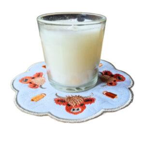 Round embroidered Highland cow candle mat in white with four cow faces arranged in a circular design, shown beneath a small glass jar candle.
