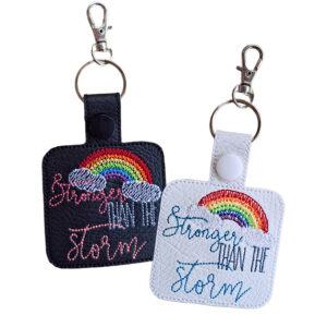 Two rectangular keyrings embroidered with the phrase “Stronger than the storm” beneath a colourful rainbow and white clouds. One keyring is black with pink and grey embroidery, and the other is white with blue and grey embroidery. Both have a snap button, silver split ring, and lobster clasp