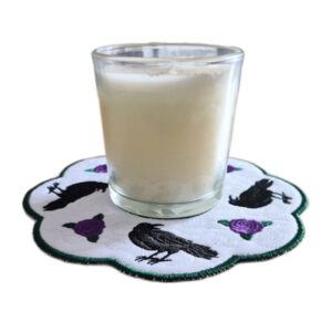 Embroidered raven candle mat with scalloped edge, featuring black ravens and rose accents on a white background, holding a glass candle on top.