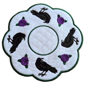 Close-up of an embroidered raven candle mat with scalloped edge, featuring detailed black ravens and rose accents arranged around a central circle on a textured white background.