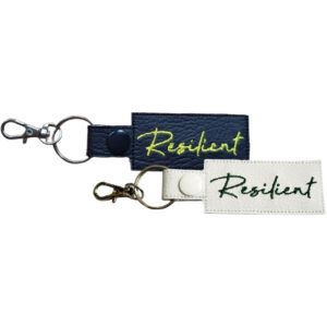 Two rectangular keyrings with the word "Resilient" embroidered in cursive. One keyring is black with yellow embroidery, and the other is white with dark green embroidery. Both feature silver keyrings and lobster clasps.