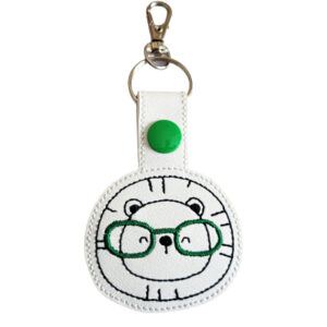Roarsome Lion with Glasses Keyring featuring an embroidered lion face wearing green glasses on white faux leather, with a green snap button and silver clasp.
