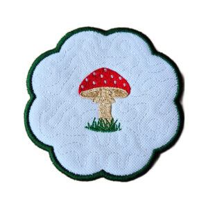 Round scalloped candle coaster featuring a single embroidered toadstool at the centre, set on a stitched background with a contrasting green border.