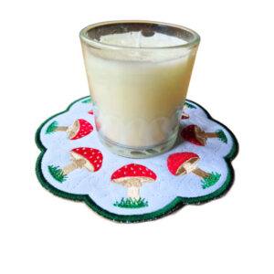 Round scalloped candle mat featuring embroidered toadstools arranged in a circular design, shown beneath a small glass jar candle.