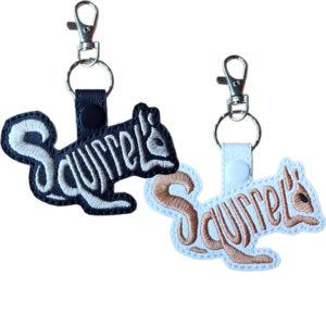 Two faux leather squirrel word art keyrings, one in black with white embroidery and one in white with brown embroidery, each featuring the word “Squirrel” forming the animal’s shape and finished with a metal clasp and snap fastening.