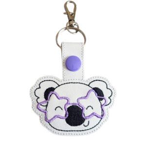 Starry-Eyed Koala with Glasses Keyring featuring an embroidered koala face wearing star-shaped glasses on white faux leather, with a purple snap button and silver clasp.