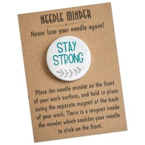 Stay Strong Needle Minder with teal script text and a small grey branch design on a round white 38mm magnet, used to hold needles for cross stitch or embroidery, displayed on kraft card packaging with instructions.