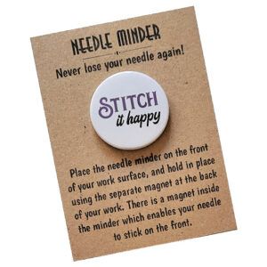 Circular needle minder with white background and colourful text that reads ‘Stitch It Happy’ in playful lettering. The design is mounted on kraft card packaging with the heading ‘Needle Minder – Never lose your needle again!’ and usage notes.