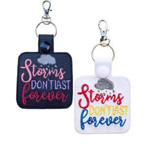 Two rectangular keyrings embroidered with the phrase “Storms don’t last forever” and a small raincloud above the text. One keyring is black with pink and purple embroidery, and the other is white with red, yellow, and blue embroidery. Both have a snap button, split ring, and lobster clasp.